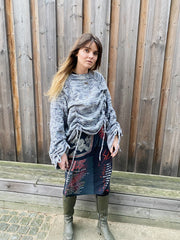 Woman wearing a gray sweater and patterned skirt standing against a wooden fence.
