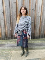 Woman wearing a gray sweater and colorful skirt standing against a wooden fence.